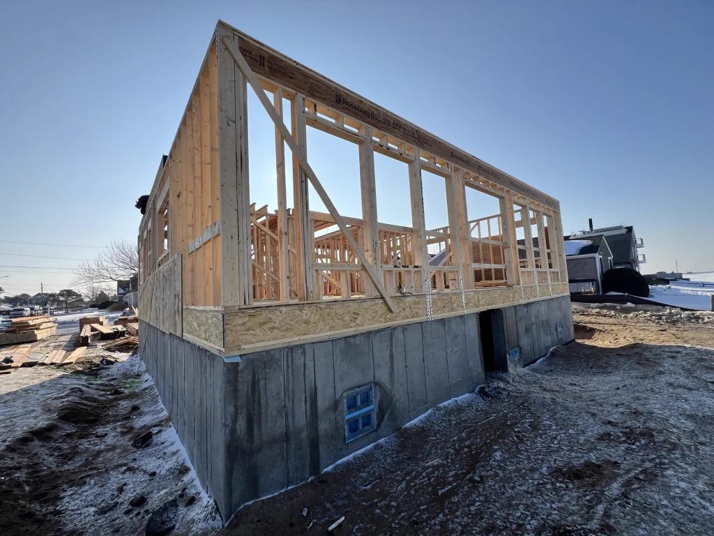 Walls and engineered beams framed on the elevated foundation, water visible behind